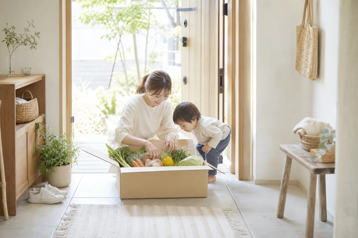 玄関で届いた食材の箱を開けるママと子ども