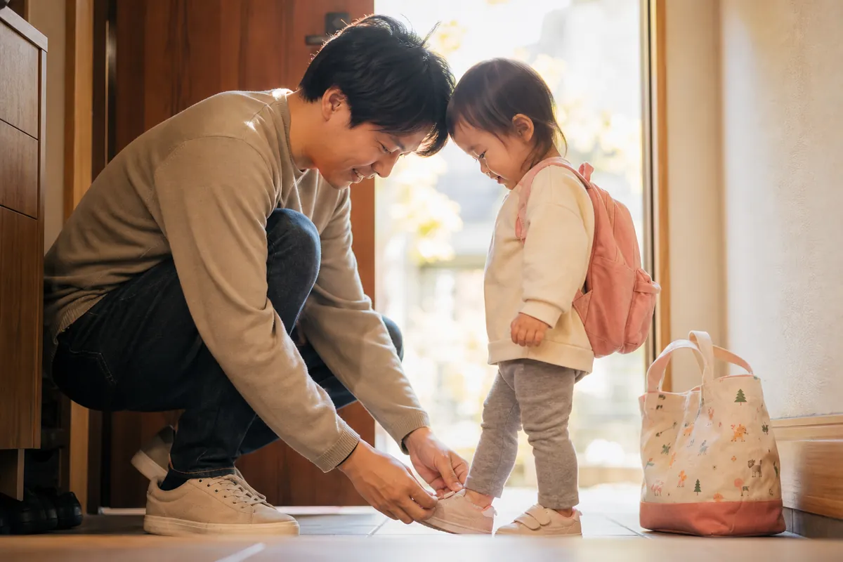 朝の玄関で子どもに靴をはかせて登園の練習をするパパ